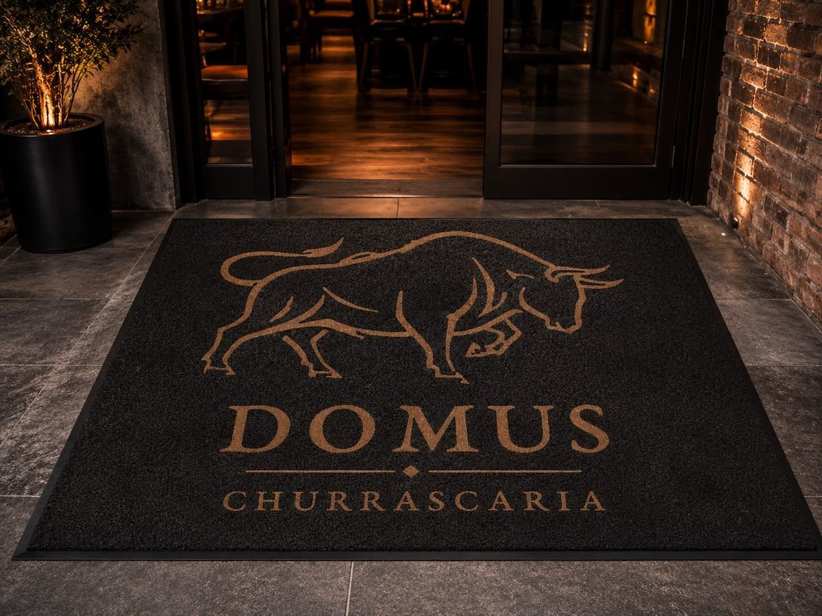Black entrance mat with gold bull logo and text reading Domus Churrascaria at restaurant entrance