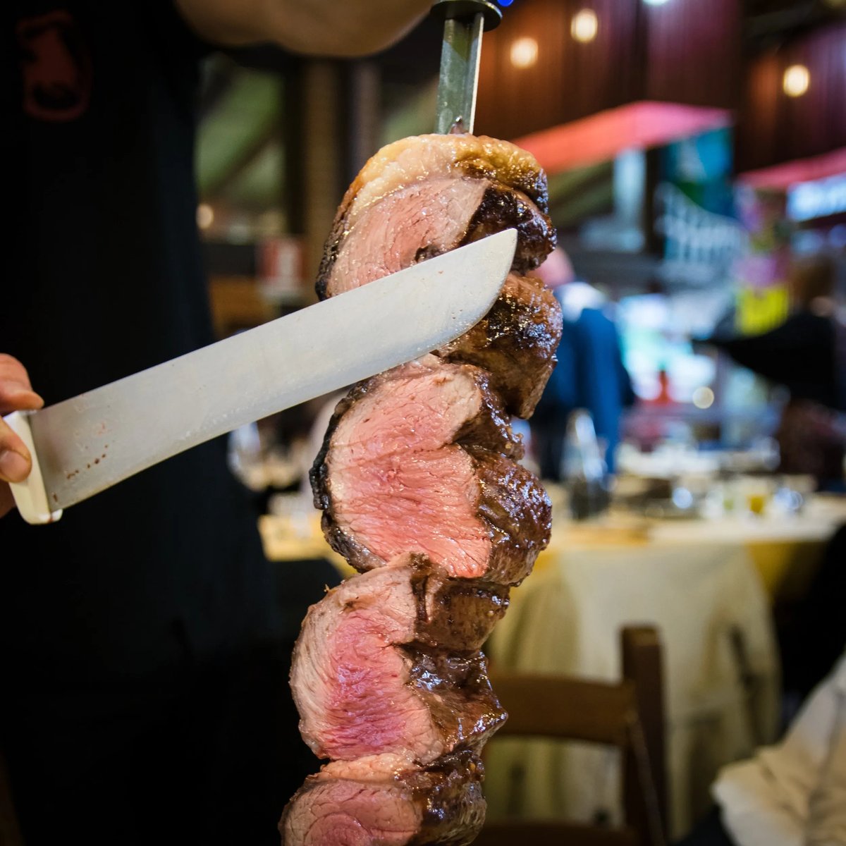 A knife cuts through stacked slices of medium-rare beef on a skewer at DOMUS Churrascaria