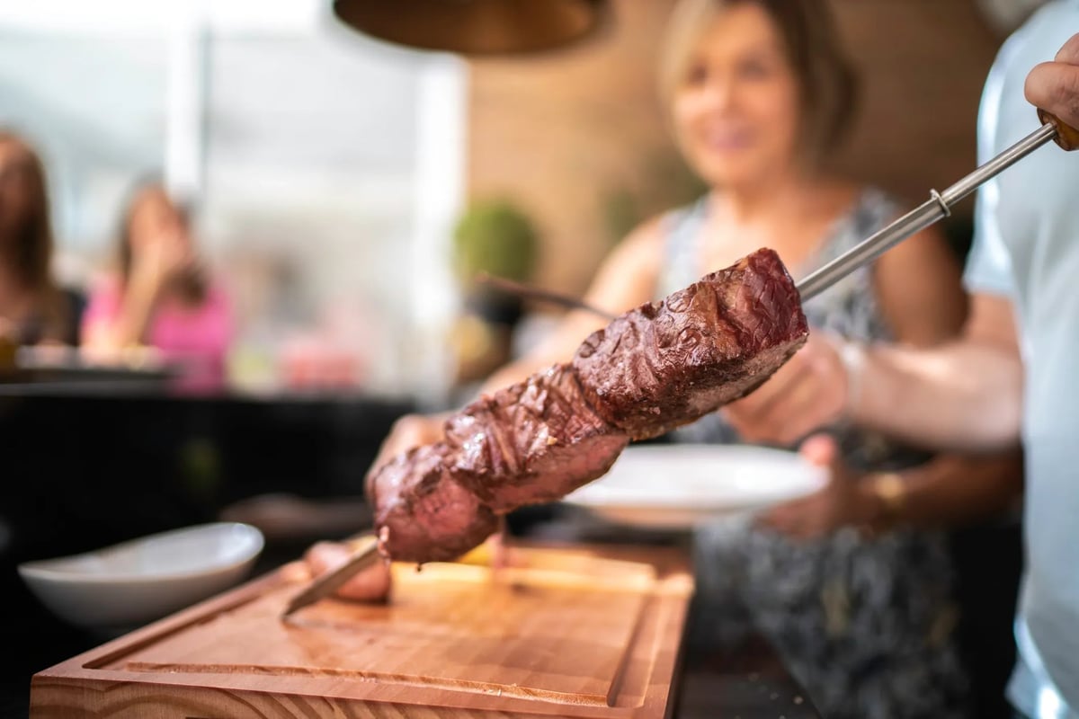 Grilled meat skewer held over a wooden cutting board by the passador at DOMUS Churrascaria
